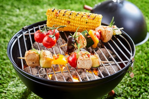 Vegetables and tofu kebab grilling on grid grille with fresh corn, viewed in close-up against green lawn grass in background