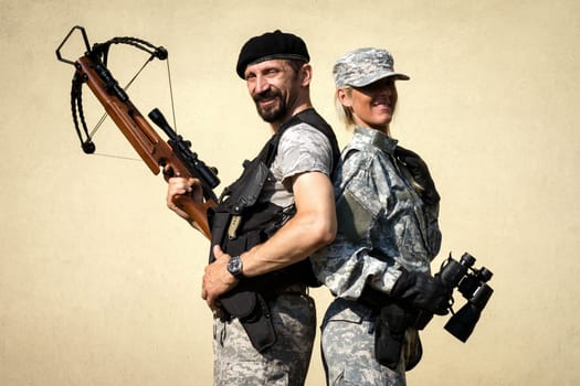 The smiling male and female soldiers in military uniform is posing with crossbow weapon and binoculars.
