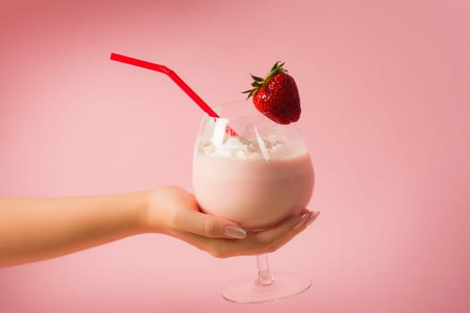 Female hand is holding a delicious healthy strawberry milkshake drink with whipped cream on bright pink background.