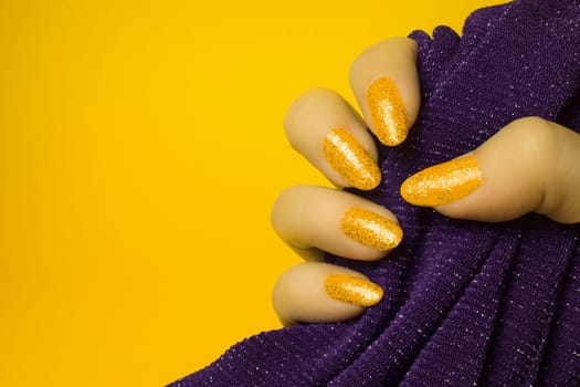 Female hand with glittered yellow nails is holding purple textile on yellow background, manicure and nail care concept.