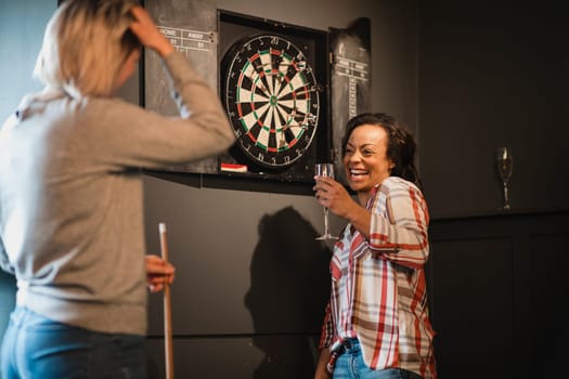 Two female friends relaxing and having fun. They are standing in a games room next to a darts board, enjoying a drink.