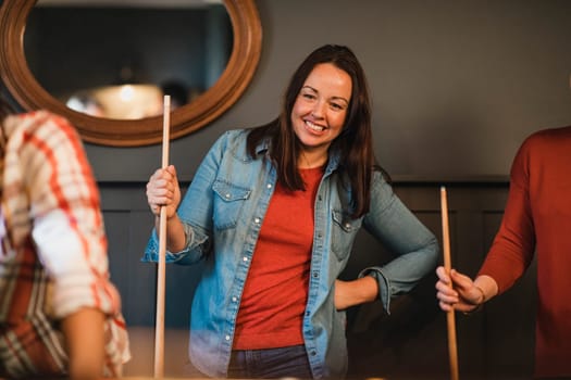 Mid adult woman standing indoors while playing a game of pool. She is holding a pool cue and watching the game play out while waiting for her go.