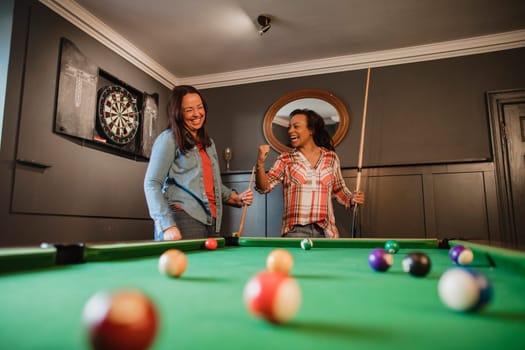 Two mid adult friends playing a game of pool in a games room in a house.