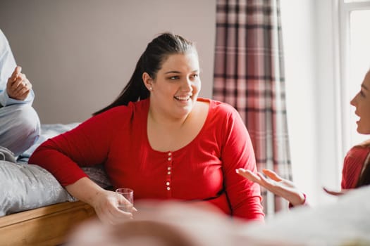Young female adult sitting in a bedroom in her pyjamas while talking to her friends. She is sitting smiling and enjoying a glass of champagne.