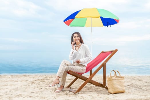 Asian business woman talking on mobile phone while sitting sitting in the beach chair with colorful umbrella on beach. Summer vacation