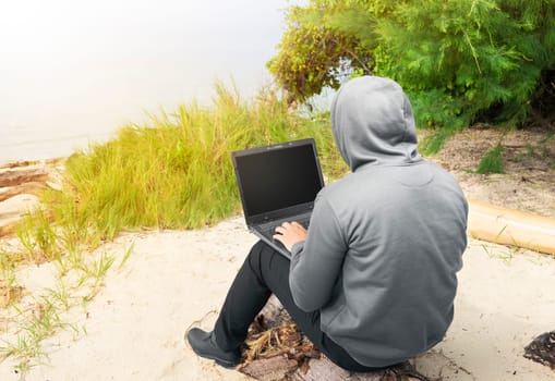 Rear view of man in black hoodie using laptop on the beach