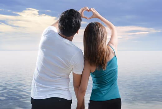 Rear view of asian romantic couple making heart shapes with fingers on the beach. Summer vacation