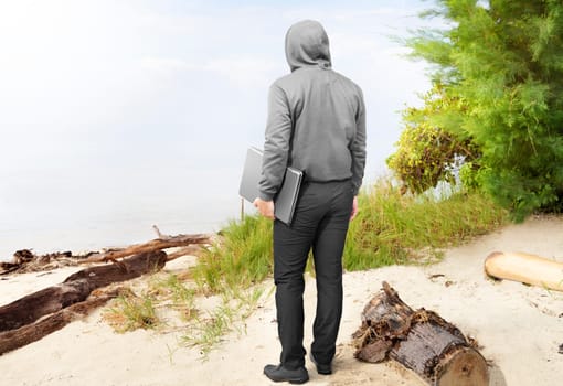 Rear view of man in black hoodie carrying laptop in his hand on the beach