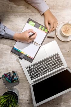 Top view of businessman writing in notepad with laptop, coffee, potted plant and business accessories on the desk
