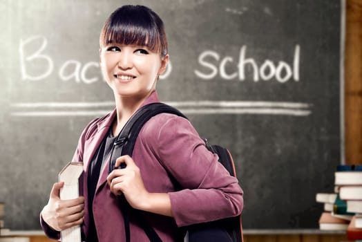 Asian student woman with backpack carrying book standing and look back in the classroom with piles of books and blackboard background. Back to School concept