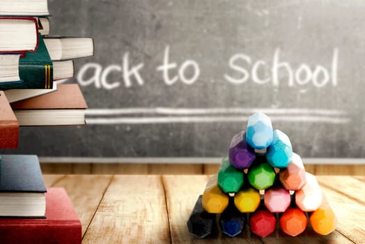 Close up view of pile of books and colorful crayons on wooden table and blackboard with Back to School message. Back to School concept