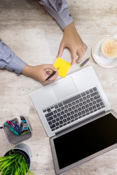 Top view of businessman writing in sticky notes with laptop, coffee, potted plant and business accessories on the desk