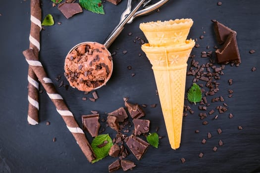 Chocolate ice cream in scoop with wafer sticks, cone and chocolate on a black slate board. Focus on ice cream in scoop and on wafer cone.