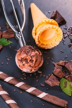 Chocolate ice cream in scoop with wafer sticks, cone and chocolate on a black slate board. Focus on ice cream in scoop.