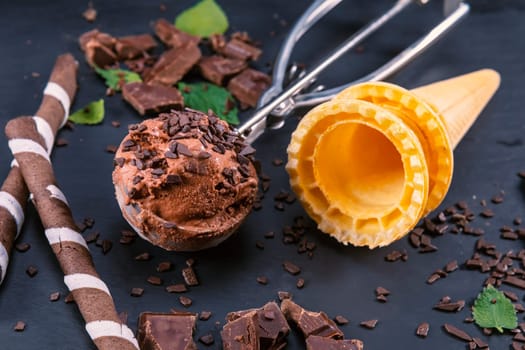 Chocolate ice cream in scoop with wafer sticks, cone and chocolate on a black slate board. Focus on ice cream in scoop.