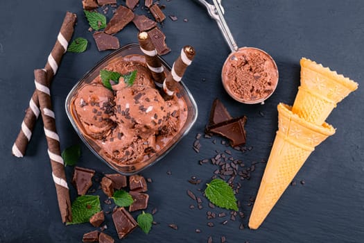 Scoops chocolate ice cream in glass bowl with wafer sticks, cone and chocolate on a black slate board. Focus on Bowl with scoops chocolate ice cream