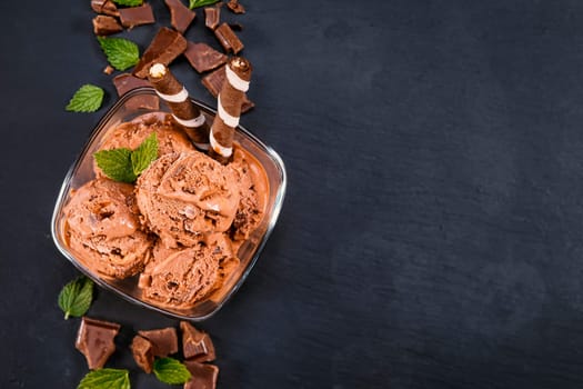Scoops chocolate ice cream in glass bowl with wafer sticks, cone and chocolate on a black slate board. Focus on Bowl with scoops chocolate ice cream