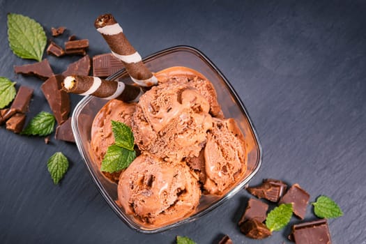 Scoops chocolate ice cream in glass bowl with wafer sticks, cone and chocolate on a black slate board. Focus on Bowl with scoops chocolate ice cream.