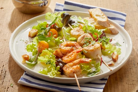 Tasty grilled spicy seasoned fresh scampi and scallop on kebab skewers served with leafy greens, baguette slices and fried croutons in a close up view