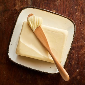 Pat of farm butter with wooden spreader with a scoop of butter on top viewed from above on a rustic wood table in square format