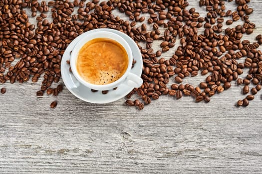 Cup of delicious hot frothy espresso coffee with scattered brown roasted beans viewed top down on rustic weathered wood with copy space below