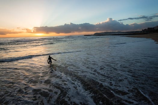 Surfer silhouetted dawn sunrise going surfing beach entry into ocean wave landscape.