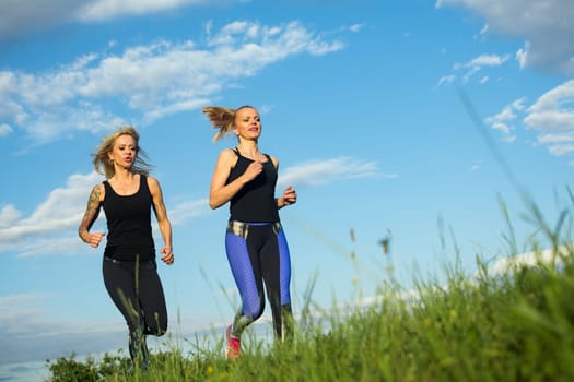 Two blond hair sporty women are jogging outdoors in nature on blue sky background.
