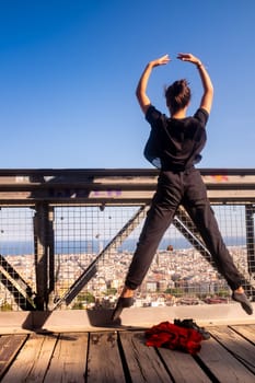 Elegant ballet performance by a young female dancer wearing a black dress, she is jumping in mid-air in front of a panoramic view of Barcelona, Spain.