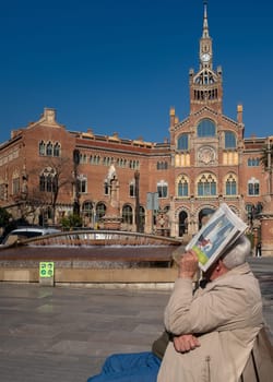 An elderly man is sitting on a bench and covers his face with a newspaper, he seems to look towards the main facade of the Hospital of the Holy Cross and St. Paul, Barcelona, Spain