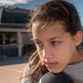 Beautiful teenage girl with long brown hair and cerulean eyes, thoughtful looking elsewhere, sitting relaxed outside, blurred background of a building.