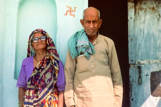 Portrait of a indian couple in old age, traditional clothing, standing at the door in front of their house, religious symbol painted on the wall, India