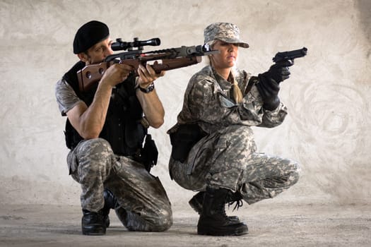 The male and female soldiers in military uniform are aiming with crossbow and pistol and squatting on gray wall background.