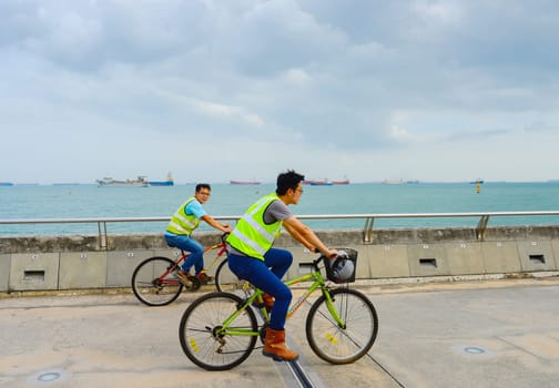 SINGAPORE - FEBRUARY 16, 2107: Two worker in uniform riding the bicycles at Singapore harbor. Industrial cargo ships in the background