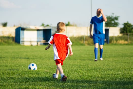 Little soccer boy in red and trainer in blue soccer dress are playing soccer on playing field, soccer training for children concept.