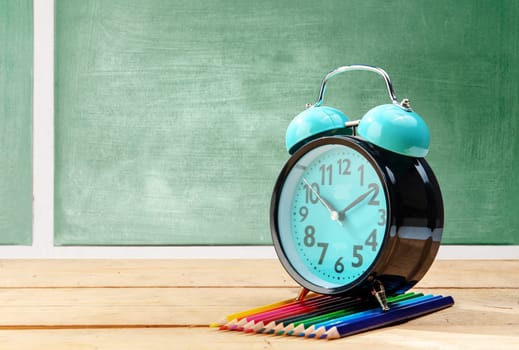 Alarm clock and colored pencils on wooden table with chalkboard background. Back to School concept