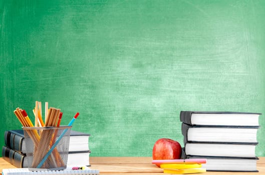 Pile of books with notes paper and pen, apple and pencils in basket container on wooden table with chalkboard background. Back to School concept