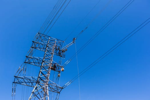 Electricians unidentified men high up suspended on electrical steel tower rigging installing new high voltage electricity power-lines.