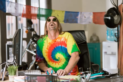 L:aughing man in tie dye shirt and round glasses at his desk