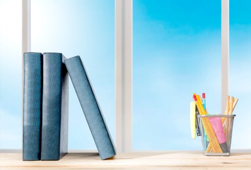 Pile of books standing and pencils in basket container with green stapler on the wooden table with window glass background. Back to School concept