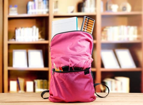 Pink backpack with book and different stationery on the wooden table in library. Back to School concept