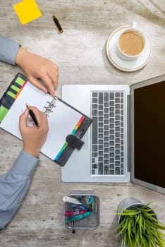 Top view of businessman writing in notebook with laptop, coffee, sticky notes, potted plant and business accessories on the desk