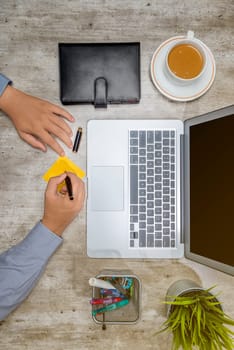 Top view of businessman writing in sticky notes with laptop, coffee, notebook, potted plant and business accessories on the desk