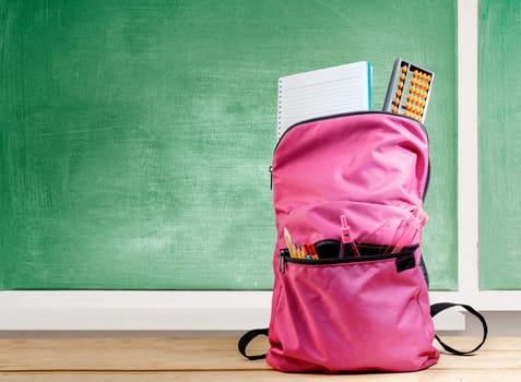 Pink backpack with book and different stationery on the wooden table with chalkboard background. Back to School concept