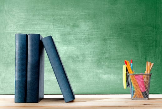 Pile of books standing and pencils in basket container with green stapler on the wooden table with chalkboard background. Back to School concept