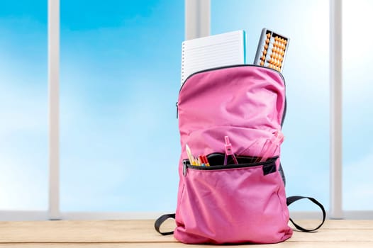 Pink backpack with book and different stationery on the wooden table with window glass background. Back to School concept