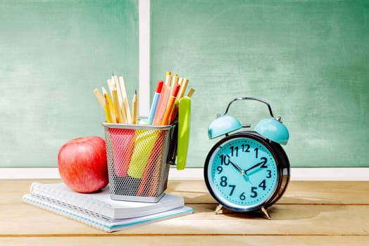 Pile of notebooks with apple and pencils in basket container with green stapler and alarm clock on the wooden table with chalkboard background. Back to School concept