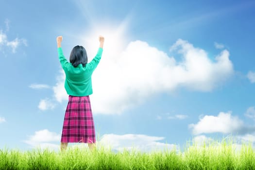 Rear view of asian cute girl standing while raised arms on the grass field with sunlight over blue sky background. Back to School concept