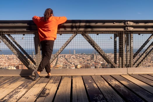 Back view of a teenager girl wearing an orange hooded shirt standing on the bridge. Relaxing pose. Contemplating the urban landscape and thinking about life, concepts.