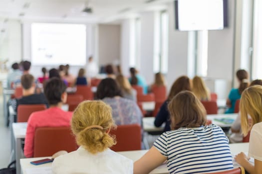 Speaker giving presentation in lecture hall at university. Participants listening to lecture and making notes.