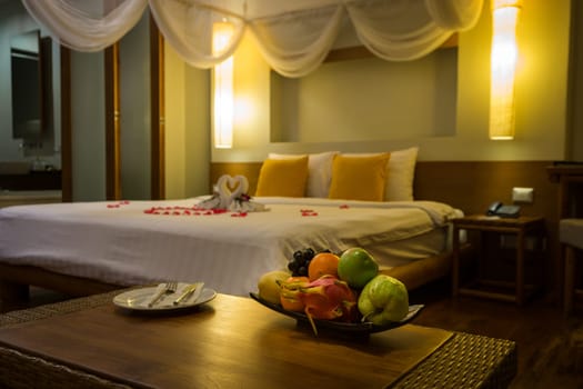 Bowl of exotic fruits and blurred background with two white towel swans, flowers and red rose petals shaped in big heart on the double bed, Honeymoon decoration and concept